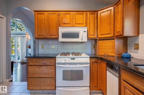 Kitchen featuring wood-finish cabinetry, dark countertops, white tile flooring, and integrated white appliances - 8 Edgemont Point(E), St. Albert, AB 
