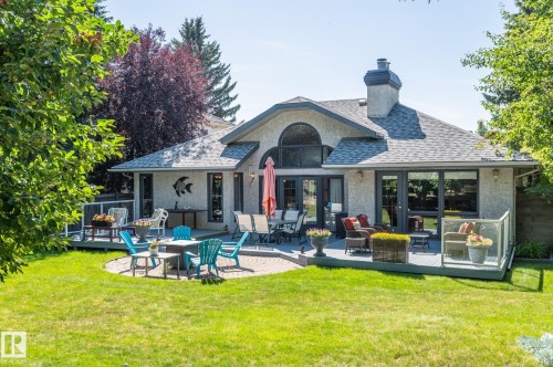Expansive backyard featuring a manicured lawn, a large deck with glass railings, a paved patio area, and a stucco exterior with a prominent arched window - 8 Edgemont Point(E), St. Albert, AB 