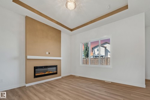 Recessed ceiling details with accent trim, built-in electric fireplace with mantel, window with white trim, wood-finish flooring, and a decorative ceiling light fixture - 32 Dalquist Bay, Leduc, AB 