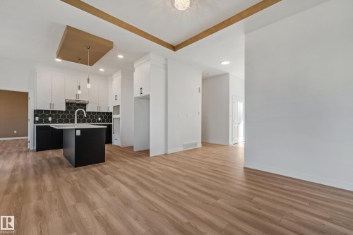 Open concept living area featuring wood-finish flooring, a kitchen island with sink, a black hexagonal tile backsplash, white upper cabinetry, and a gold-accented ceiling - 32 Dalquist Bay, Leduc, AB 
