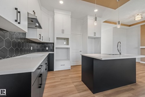 Kitchen featuring wood-finish flooring, two-tone cabinetry, geometric tile backsplash, light-toned countertops, and a center island with a built-in sink - 32 Dalquist Bay, Leduc, AB 