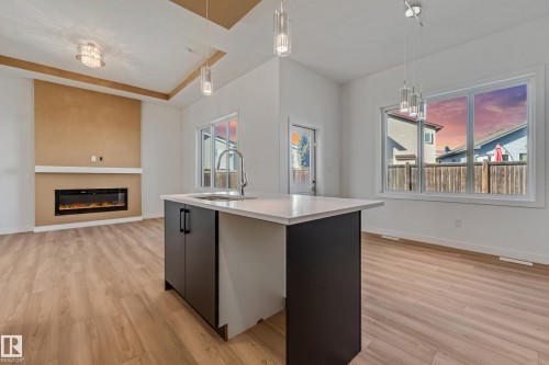 Central island featuring a stainless steel sink and matte black cabinetry, wood-finish flooring, and a modern fireplace with a mantel - 32 Dalquist Bay, Leduc, AB 