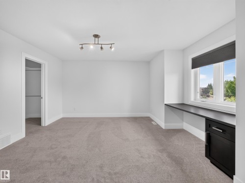 Spacious room featuring neutral carpeting, white walls, a built-in dark wood desk with a single cabinet, a large window, and a track lighting fixture - #59 10550 Ellerslie Rd Sw, Edmonton, AB - Indoor Photo Showing Other Room