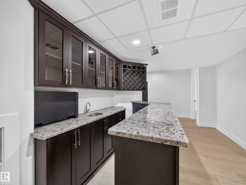 Wet bar featuring granite countertops, a built-in sink, dark wood-finish cabinetry with glass panel doors, and integrated wine storage - #59 10550 Ellerslie Rd Sw, Edmonton, AB - Indoor Photo Showing Other Room