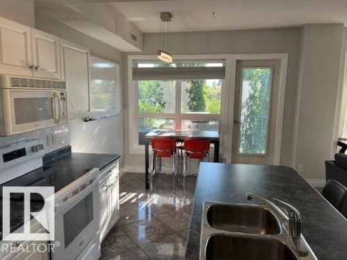 White cabinetry with silver hardware, dark countertops, built-in microwave, white electric range, and a double basin stainless steel sink with a chrome faucet - #407 11103 84 Av Nw, Edmonton, AB 