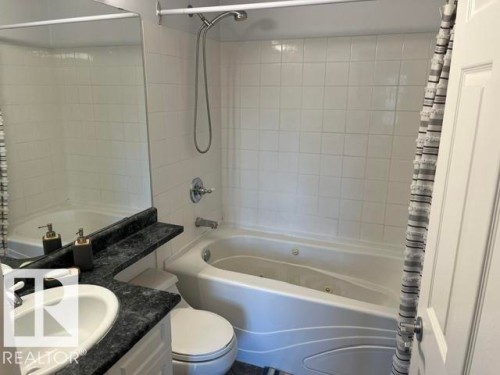 Corner soaking tub with integrated jets, white subway tile surround, polished chrome fixtures, and a dark stone-finish vanity with an undermount sink - #407 11103 84 Av Nw, Edmonton, AB 