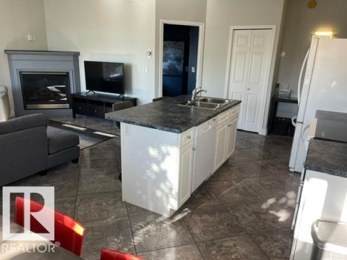 Tile flooring throughout, kitchen island with integrated sink, white cabinetry, dark countertops, and a fireplace with a gray mantel - #407 11103 84 Av Nw, Edmonton, AB 