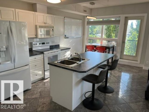Kitchen featuring white cabinetry, white appliances, dark countertops, a center island with an integrated sink, and tile flooring - #407 11103 84 Av Nw, Edmonton, AB 