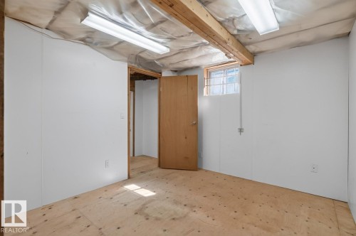Unfinished room featuring white panel walls, exposed ceiling joists, two fluorescent light fixtures, a light wood door, and a single window - 12309 128 Street Nw, Edmonton, AB 
