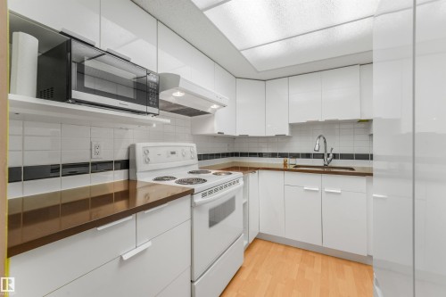 White cabinetry with modern pulls, white tile backsplash with black accent, dark countertops, and wood-finish flooring - #203 10171 119 St Nw, Edmonton, AB 