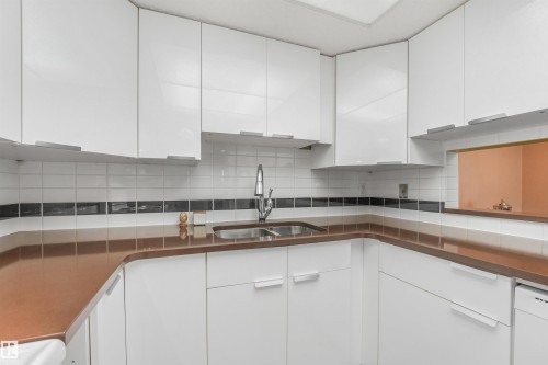 White flat-panel cabinetry, stainless steel double basin sink, chrome gooseneck faucet, brown solid surface countertops, and white subway tile backsplash with accent band - #203 10171 119 St Nw, Edmonton, AB 