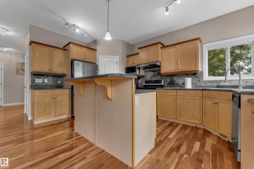 Wood-finish flooring, light wood cabinetry with dark hardware, dark countertops, and stainless steel appliances - 1226 Mcallister Way Sw, Edmonton, AB 