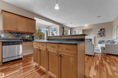 Wood-finish flooring throughout, wood cabinetry with dark countertops, stainless steel dishwasher, and a stone tile backsplash - 1226 Mcallister Way Sw, Edmonton, AB 