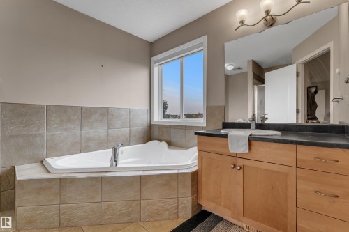 Alcove soaking tub with tile surround, wood-finish vanity with dark countertop, integrated sink, and an expansive wall-mounted mirror - 1226 Mcallister Way Sw, Edmonton, AB 