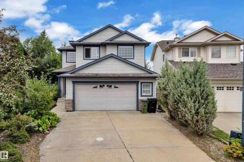 Two-story exterior with light gray siding and blue trim, featuring a two-car garage, concrete driveway, and mature landscaping - 1226 Mcallister Way Sw, Edmonton, AB 