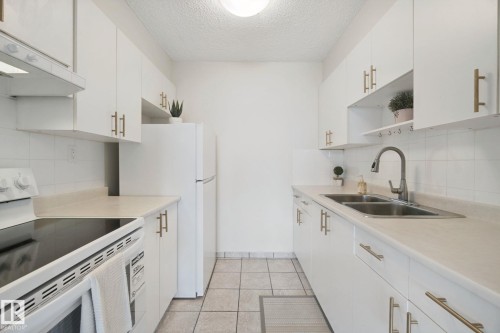 White cabinetry with gold-tone hardware, white tile backsplash, dual basin stainless steel sink, and light-toned countertops - #104 8745 165 St Nw, Edmonton, AB 