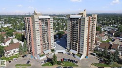 Two high-rise buildings featuring multiple balconies, surrounded by paved parking areas and mature trees - 