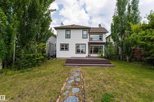 White siding exterior with dark trim windows, multi-level wood-finish deck, stone pathway, and mature perimeter trees - 5092 Thibault Way Nw, Edmonton, AB 