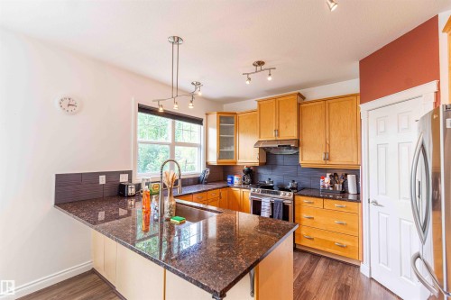 Kitchen featuring stainless steel appliances, wood-finish flooring, a dark tile backsplash, dark countertops, and light wood cabinetry - 5092 Thibault Way Nw, Edmonton, AB 