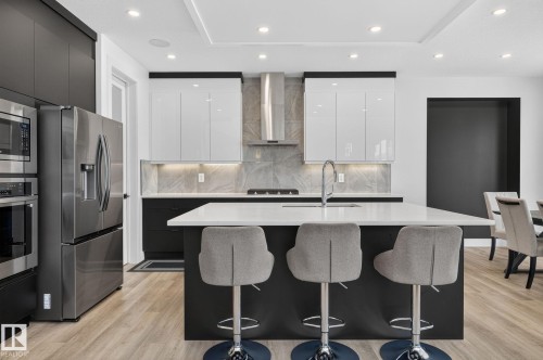 Contemporary kitchen featuring a large central island with seating, stainless steel appliances, gloss white upper cabinetry, matte black lower cabinetry, and wood-finish flooring - 16304 19 Avenue Sw, Edmonton, AB 