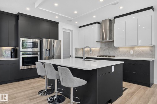 Modern kitchen featuring a central island with a white countertop and seating, stainless steel appliances, two-tone cabinetry with black base units and white upper cabinets, a stainless steel range hood, and light wood-finish flooring - 16304 19 Avenue Sw, Edmonton, AB 