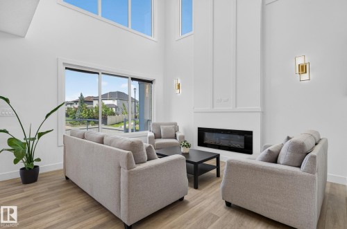 Living area featuring high ceilings, wood-finish flooring, a contemporary electric fireplace, large windows, and modern wall sconces - 16304 19 Avenue Sw, Edmonton, AB 