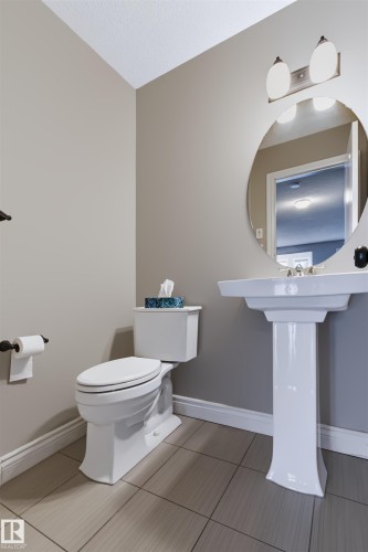 Pedestal sink with a circular mirror, wall-mounted vanity light, white toilet, and tile flooring - 1420 158 Street Sw, Edmonton, AB 
