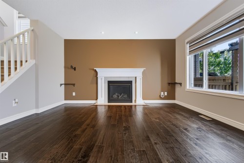 Dark wood-finish flooring throughout, featuring a fireplace with a white mantel, and a large window with roller shades - 1420 158 Street Sw, Edmonton, AB 