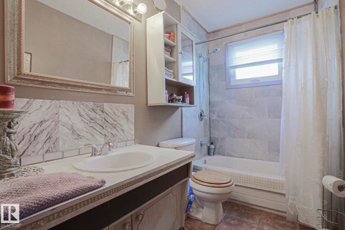 Bathroom featuring a vanity with an integrated sink, a framed wall mirror, a tiled shower/tub combination, wall-mounted cabinetry, and a window with blinds - 11440 144 Avenue Nw, Edmonton, AB 