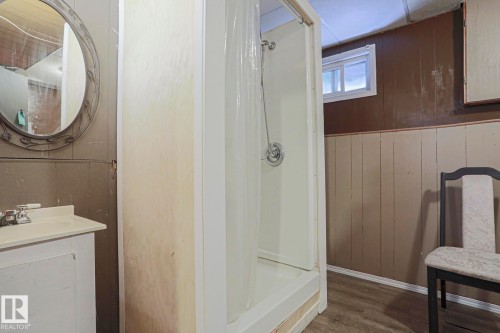 Bathroom featuring a walk-in shower enclosure with glass surround, pedestal sink with chrome fixtures, circular illuminated mirror, wood-finish flooring, and paneled wainscoting with neutral tile detailing - 11440 144 Avenue Nw, Edmonton, AB 