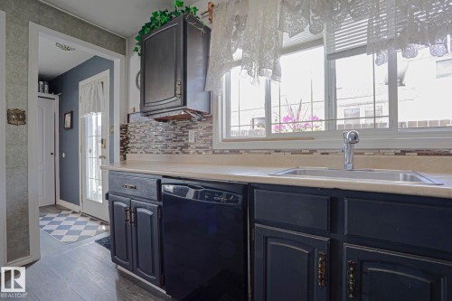 Kitchen featuring dark cabinetry, light-toned countertops, a stainless steel sink with a chrome faucet, a tile backsplash, and wood-finish flooring - 11440 144 Avenue Nw, Edmonton, AB 