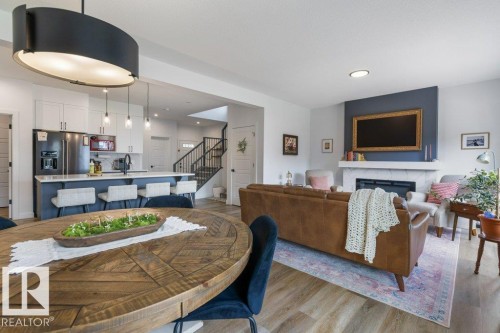 Open-concept living space featuring wood-finish flooring, a kitchen island with seating, stainless steel appliances, a fireplace with a mantel, and a black wrought iron staircase - 34 Raspberry Road, St. Albert, AB 