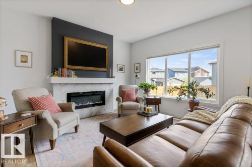 Living room featuring a large picture window, wood-finish flooring, and a modern fireplace with a white mantel and light-toned surround - 34 Raspberry Road, St. Albert, AB 