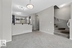 Light gray wall finishes and carpeting throughout, featuring a breakfast bar with dark countertops, a staircase with a white banister, and ceiling-mounted light fixtures - 