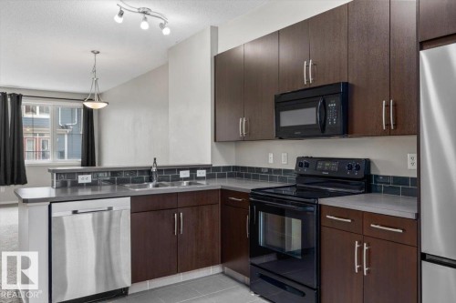 Modern kitchen featuring dark wood-finish cabinetry, stainless steel appliances, and a tiled backsplash - #31 603 Watt Bv Sw, Edmonton, AB - Indoor Photo Showing Kitchen With Double Sink