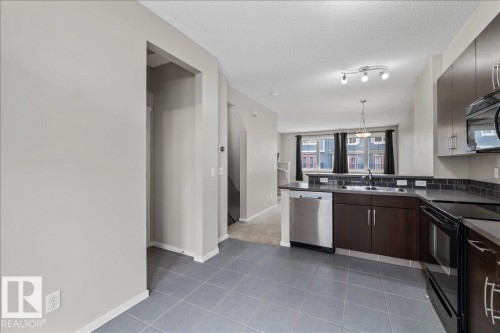 Kitchen featuring dark wood-finish cabinetry, stainless steel dishwasher, black oven, tile backsplash, and light gray tile flooring - #31 603 Watt Bv Sw, Edmonton, AB - Indoor Photo Showing Kitchen With Double Sink
