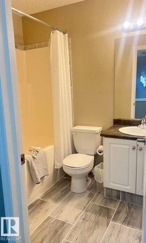Bathroom featuring wood-finish tile flooring, a shower-over-tub combination with a tiled surround, a white toilet, and a vanity with a white basin and dark countertop - 5001 62 Street, Beaumont, AB 