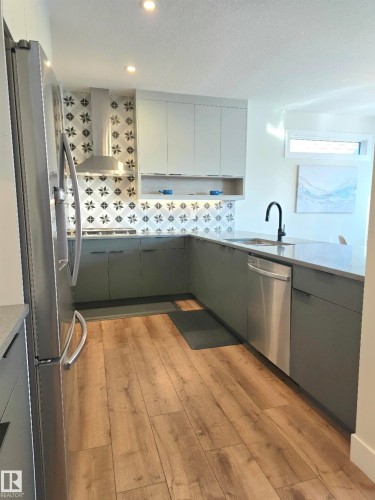 Modern kitchen featuring stainless steel appliances, sleek gray cabinetry with white upper units, patterned tile backsplash, black matte faucet, and warm wood-finish flooring throughout the galley layout - 20827 130 Avenue Nw, Edmonton, AB 