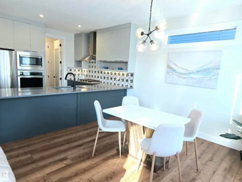 Contemporary kitchen featuring stainless steel appliances, a patterned tile backsplash, wood-finish flooring, a kitchen island with a built-in sink, and a modern light fixture - 20827 130 Avenue Nw, Edmonton, AB 