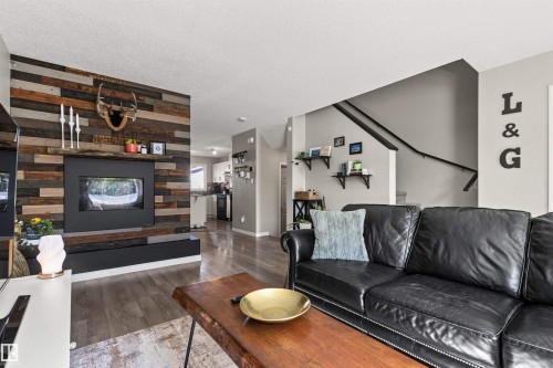 Wood-finish flooring throughout, integrated fireplace with wood panel accent wall, natural wood mantel, and a staircase with a dark handrail - 4306 Prowse Link Link Sw, Edmonton, AB 