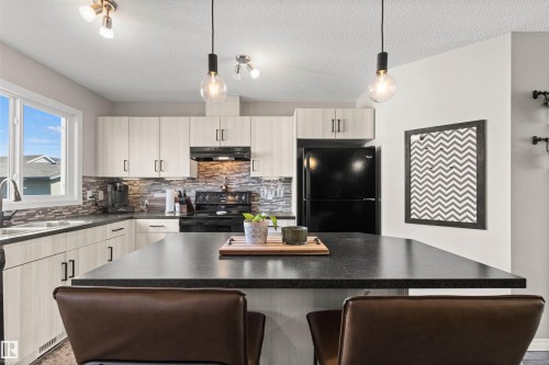 Kitchen featuring light-finish cabinetry, dark countertops, a stone-finish tile backsplash, and a central island with pendant lighting - 4306 Prowse Link Link Sw, Edmonton, AB 
