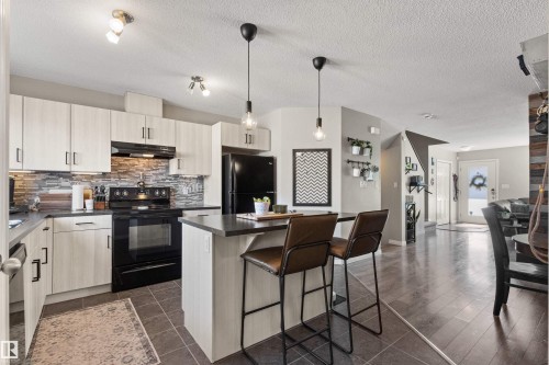 Kitchen featuring light wood-finish cabinetry, dark countertops, a stone-look tile backsplash, and black appliances - 4306 Prowse Link Link Sw, Edmonton, AB 