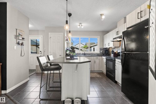 Kitchen featuring light wood-finish cabinetry, a dark tile backsplash, dark-toned flooring, a peninsula island with a dark countertop, and black appliances - 4306 Prowse Link Link Sw, Edmonton, AB 