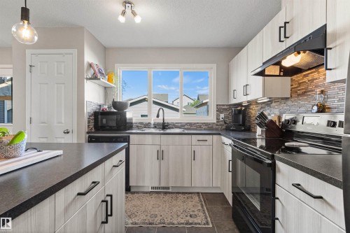 Contemporary kitchen featuring wood-finish cabinetry, dark countertops, a tiled backsplash, and stainless steel appliances - 4306 Prowse Link Link Sw, Edmonton, AB 