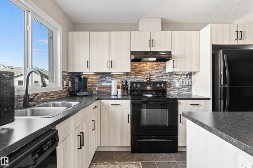 Updated kitchen featuring light wood-finish cabinetry, dark countertops, black appliances, and a mosaic tile backsplash - 4306 Prowse Link Link Sw, Edmonton, AB 
