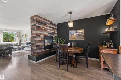 Wood-finish flooring throughout, featuring a modern fireplace with a textured wood surround, and a clear glass pendant light fixture - 4306 Prowse Link Link Sw, Edmonton, AB 