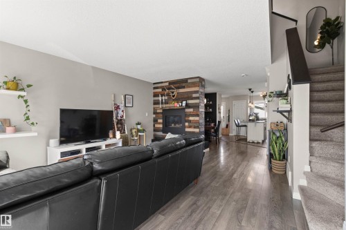 Wood-finish flooring throughout, modern fireplace with a wood-panel accent wall, built-in shelving, and carpeted stairs with a dark handrail - 4306 Prowse Link Link Sw, Edmonton, AB 