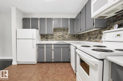 Kitchen featuring L-shaped counter space, gray cabinetry, white appliances, and a tiled backsplash - #203 2624 Mill Woods Rd E Nw, Edmonton, AB 