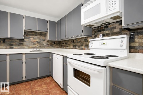 Kitchen featuring gray cabinetry, light-toned countertops, a multi-tone tile backsplash, and a white oven/stove combination - #203 2624 Mill Woods Rd E Nw, Edmonton, AB 