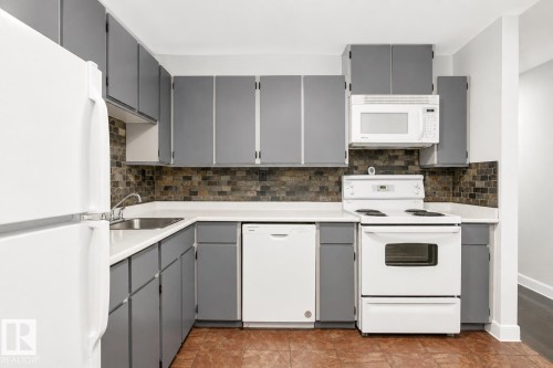 Efficient galley kitchen featuring gray cabinetry with chrome hardware, white appliances including electric range and microwave, tile backsplash, laminate countertops, and wood-finish flooring throughout - #203 2624 Mill Woods Rd E Nw, Edmonton, AB 